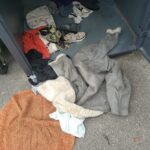 Atvērts metāla apģērbu savākšanas konteiners, no kura izbirusi daudzveidīga apģērbu kaudze, tostarp pelēks aitas vilnas mētelis un oranžs paklājs uz zemes.
