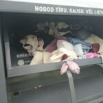 Pelēks tekstila savākšanas konteiners, kura atvērtā augšdaļa ir pilna ar dažādu krāsu drēbēm. Uz konteinera rakstīts: "NODOD TĪRU. SAUSU. VĒL LIETOJAMU TEKSTILU." un "DRĪKST APĢĒRBU" blakus T-krekla ikonai.