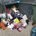 Pelēka tekstila savākšanas tvertne ir pārpildīta, un drēbes kopā ar citiem tekstilizstrādājumiem ir izbirušas uz bruģa. Blakus atrodas zaļa atkritumu tvertne. Uz pelēkās tvertnes ir uzraksts "MĀJAS TEKSTILS". Uz vienas baltas somas uz zemes redzami vārdi "paldies!", "dēkuju!", "thanks!", "aitäh!", bet uz citas somas "pepo". Uz zaļās tvertnes ir uzraksti "MEST", "NEMEST", "LAI LABI".