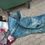 Netīra, zila stepēta sega un kaudze drēbju, tostarp balts maiss ar uzrakstu "pepco", guļ uz bruģa pie zaļa atkritumu konteinera.