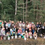 Pieaugušo grupa sapulcējusies uz zāles pie bērnu rotaļu laukuma, ar lielu koku un ēku fonā, rudens dienā.