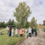 Uz zaļa zāliena, blakus bērnu rotaļu laukumam un ēkai, apmēram desmit pieaugušo stāv puslokā, skatoties uz lielu bērzu. Debesis ir apmākušās.