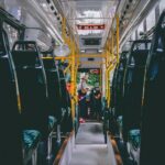 Autobusa salons ar rindām tumšu sēdvietu un dzelteniem rokturiem. Autobusa priekšgalā sieviete gaišā cepurē stāv, mijiedarbojoties ar iekārtu pie autovadītāja sēdvietas. Virs viņas redzams numurs "2166".