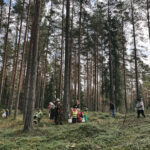 Pieaugušo grupa sapulcējusies uz zāliena pie asfaltēta celiņa, skatās virzienā uz lielu koku un bērnu rotaļu laukumu. Pa labi redzama ēka, kreisajā pusē koki ar rudenīgām lapām. Debesis ir mākoņainas.