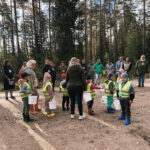 Pieaugušo grupa pulcējusies ārā, stāvot pie liela koka bērnu rotaļu laukumā. Redzams slidkalniņš, šūpoles un ēka labajā pusē.