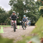 Bērni ķiverēs rindā brauc ar velosipēdiem un tricikliem pa bruģētu celiņu ar baltām pārrautām līnijām. Fonā redzama ēka.