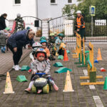 Desmit bērni krāsainās ķiverēs sarindojušies brauc ar dažādiem maziem velosipēdiem un trīsriteņiem pa bruģētu ceļu ar baltām pārtrauktām līnijām, fonā ēka.