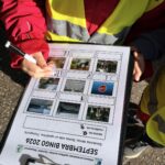 Grupa bērnu ķiverēs brauc ar dažādiem velosipēdiem un trīsriteņiem pa bruģētu celiņu ar baltām punktotām līnijām, izveidojot divas rindas, ar skolas ēku fonā.