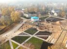 Putnu lidojuma skats uz pilsētas laukumu rudens dienā, kurā notiek rekonstrukcija. Kreisajā pusē redzama veca baznīca ar sarkanu jumtu un augstu pulksteņa torni, ko ieskauj koki ar dzeltenām lapām. Laukuma priekšplānā un centrā ir pabeigtas gājēju zonas ar celiņiem un apstādījumiem, kā arī balta, divstāvu ēka ar zilu pagaidu jumtu. Labajā pusē plaša būvlaukuma zona ar zemes kaudzēm un ekskavatoriem. Fons aizmiglots, ar citām pilsētas ēkām.