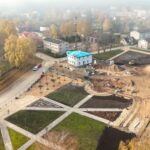 Putnu lidojuma skats uz pilsētas laukumu rudens dienā, kurā notiek rekonstrukcija. Kreisajā pusē redzama veca baznīca ar sarkanu jumtu un augstu pulksteņa torni, ko ieskauj koki ar dzeltenām lapām. Laukuma priekšplānā un centrā ir pabeigtas gājēju zonas ar celiņiem un apstādījumiem, kā arī balta, divstāvu ēka ar zilu pagaidu jumtu. Labajā pusē plaša būvlaukuma zona ar zemes kaudzēm un ekskavatoriem. Fons aizmiglots, ar citām pilsētas ēkām.