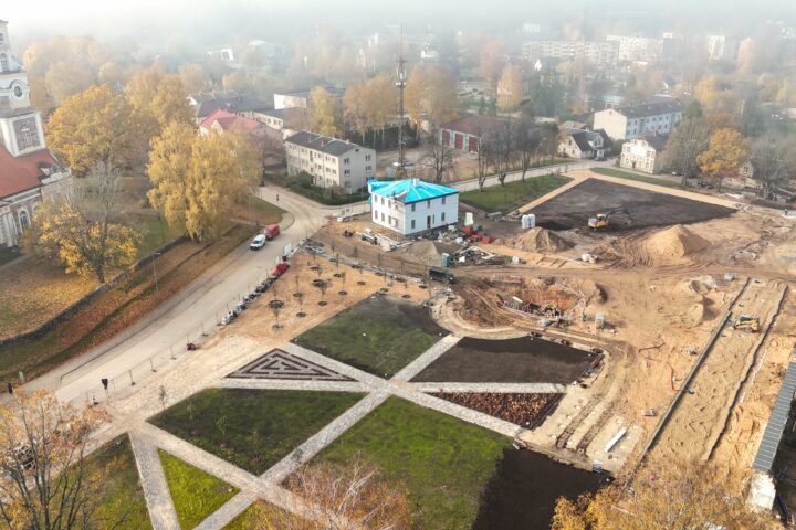 Putnu lidojuma skats uz pilsētas laukumu rudens dienā, kurā notiek rekonstrukcija. Kreisajā pusē redzama veca baznīca ar sarkanu jumtu un augstu pulksteņa torni, ko ieskauj koki ar dzeltenām lapām. Laukuma priekšplānā un centrā ir pabeigtas gājēju zonas ar celiņiem un apstādījumiem, kā arī balta, divstāvu ēka ar zilu pagaidu jumtu. Labajā pusē plaša būvlaukuma zona ar zemes kaudzēm un ekskavatoriem. Fons aizmiglots, ar citām pilsētas ēkām.