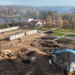 Skats no augšas uz pilsētas laukumu vai parku būvniecības procesā. Redzami bruģēti celiņi ar ģeometriskiem rakstiem un zaļi zālieni, kas mijas ar brūnām augsnes platībām un jauniem koku stādījumiem. Vienā bruģētajā zonā ir labirintveida ornaments. Laukuma malā ir iela ar automašīnām un būvmateriāliem. Apkārtnē redzamas ēkas un koki ar rudenīgām lapām.