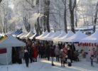 Sniegots ziemas tirdziņš ar daudzām teltīm un cilvēkiem, kas staigā starp stendiem. Fonā redzami apsarmojuši koki un balti, izgaismoti eņģeļu rotājumi. Pie viena stenda karājas sarkanas Ziemassvētku zeķes.