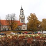Gaisa skats uz pilsētas laukumu vai parku pārbūves laikā. Bruģēti celiņi veido ģeometriskus rakstus starp zaļiem zāliena laukumiem un tumšu augsni ar iestādītiem jauniem kokiem. Gar ielu, kur stāv automašīnas, redzami būvniecības materiāli. Ielas malā redzama zīme ar burtu P. Fona redzamas ēkas un koki ar dzeltenām rudens lapām.