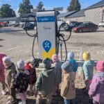 Rinda bērnu ķiverēs brauc ar dažādiem velosipēdiem un trīsriteņiem pa bruģētu ceļu, fonā redzama ēka un koki.